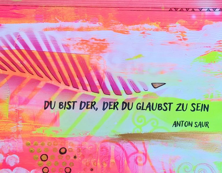 Bunte Wandmalerei mit einem Schriftzug in schwarzer Schrift, der das Zitat 'Du bist der, der du glaubst zu sein' von Anton Saur zeigt, auf einem Hintergrund aus Rosa-, Orange- und Gelbtönen mit abstrakten Mustern.